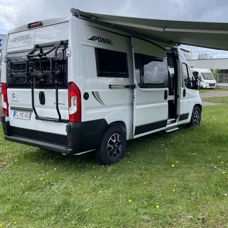 Ein grau-weißer Wohnwagen mit einer verlängerten Markise steht auf grünem Gras