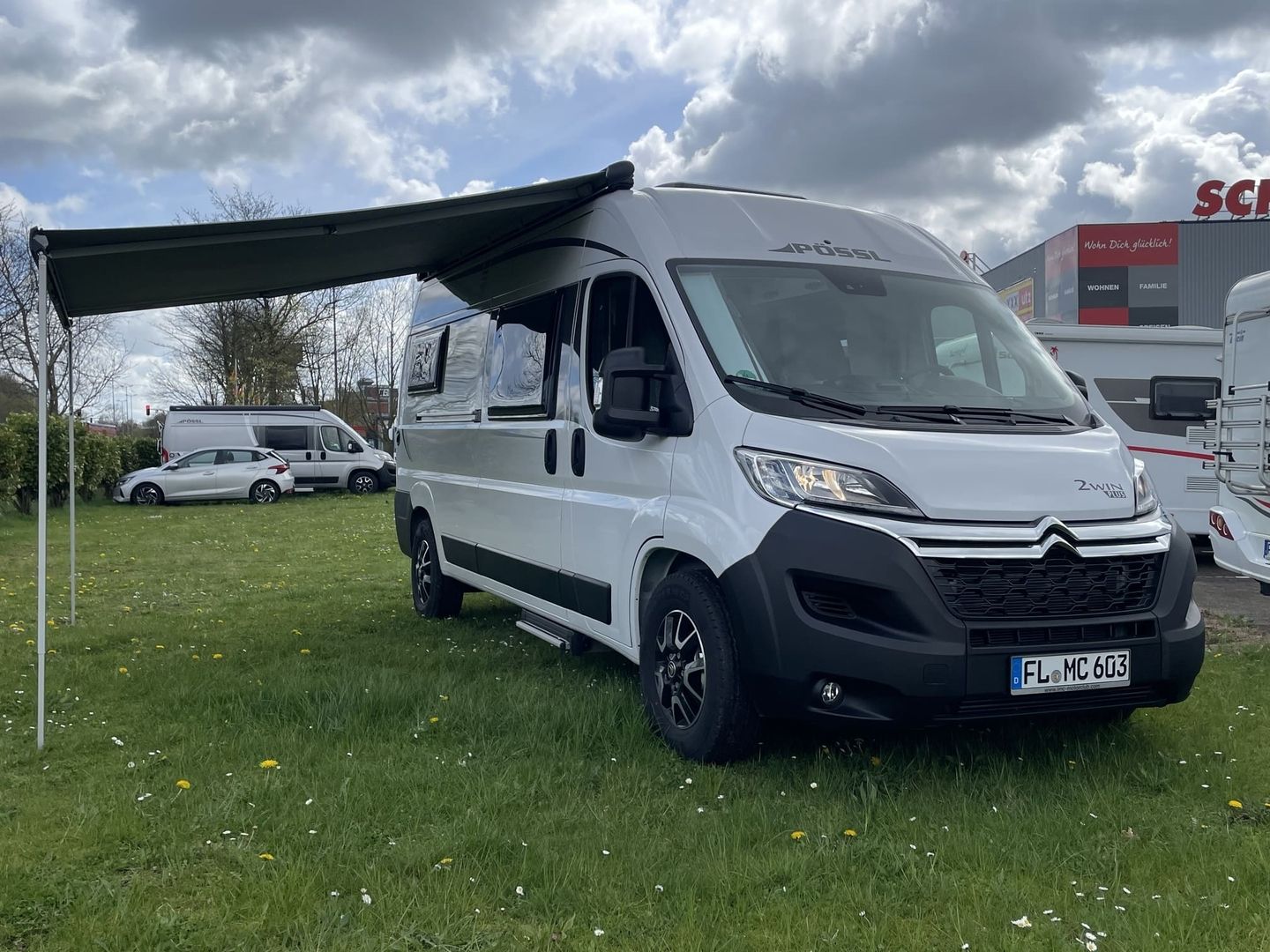 Ein grau-weißer Wohnwagen mit einer verlängerten Markise steht auf grünem Gras